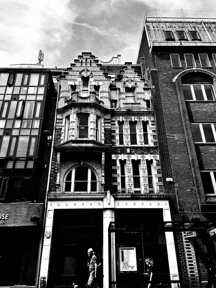 A monochrome photograph of a pair of elaborately decorated buildings with modern offices on both sides. The crow-stepped gable structures have glazed terracotta bricks with a bracketed oriel window on the second floor to the left. They date back to 1903 and are noted by Historic England as having an “exuberant mixture of Baroque Art Nouveau and Flemish styles.” 12-15 Station Road, Reading, Berkshire, UK. Photo by Gearóid Burke.