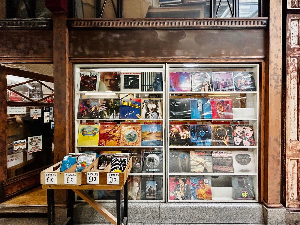 A wood-panelled shopfront with an open door and two large windows. Each window is filled shelves displaying 12" record covers in a four by five grid. Artist featured include David Bowie, The Rolling Stones, KISS, and Jimi Hendrix. A stand, just in front of the shop, offers bundles of 7" singles. The Sound Machine, 24 Harris Arcade, Reading, Berkshire, UK. thesoundmachine.uk.com. Photo by Gearóid Burke.