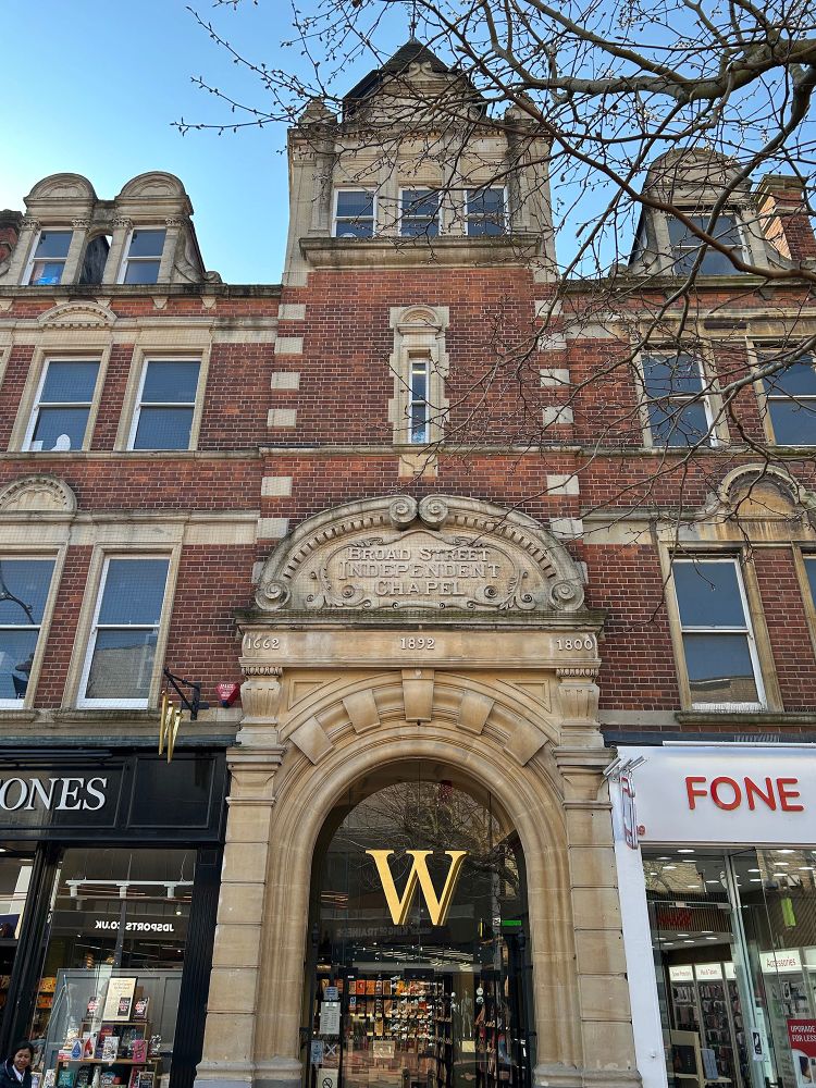 A bookshop on a British high street has an ornate arched doorway in a Victorian building. "1662 1892 1800" is inscribed in a frieze, then there is a pediment and a cartouche is inscribed "Broad Street Independent Chapel." Waterstones now occupies this frontage building and the old church behind it. 89 Broad Street, Reading, Berkshire, UK. Photo by Gearóid Burke.