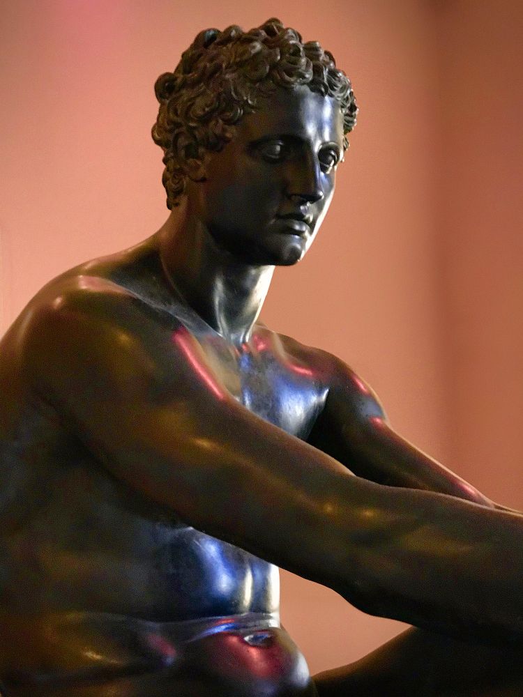 A close up of a classical bronze sculpture of a young, beardless man. The figure is seated and his arms are resting on his left knee. The background is a terracotta colour, and the mix of artificial and natural light gives the bronze blue and red highlights. Mars Seated by Luigi Valadier, bronze, 1780. The sculpture is a copy of the Ludovisi Ares, which itself is a 2nd-century copy of a late 4th-century BCE Greek original. Musée du Louvre, Paris, France. Photo by Gearóid Burke.