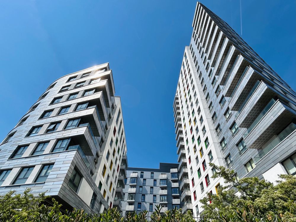 A wide-angle photograph of contemporary apartment buildings reaching up into a deep blue sky. The structures' facades are a mix of wide grey metallic cladding panels and plain white render. Some of windows have brightly-painted architraves. The Honister, Hunsaker, and Hewitt buildings, Chatham Street, Reading, Berkshire, UK. Photo by Gearóid Burke.