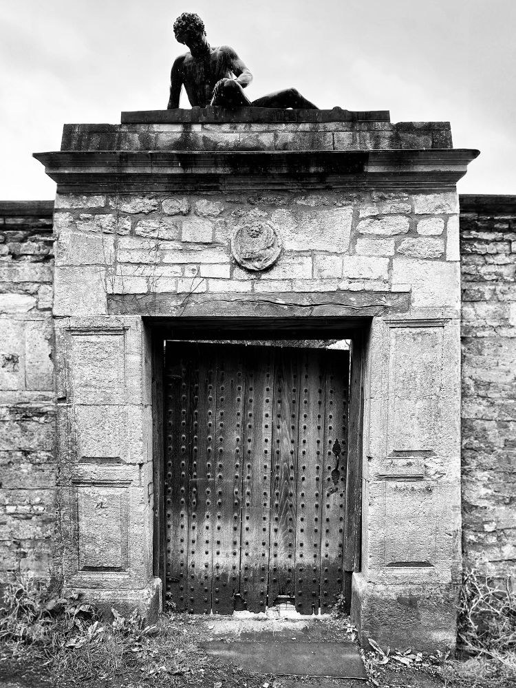 The entrance to a walled garden is overseen by a life-size, semi-recumbent bronze statue. This is a reproduction of the Dying Gaul, a Hellenistic bronze sculpture from ancient Greece that dates back to the 3rd century BCE. Iford Manor, Iford, Wiltshire, UK. Photo by Gearóid Burke.