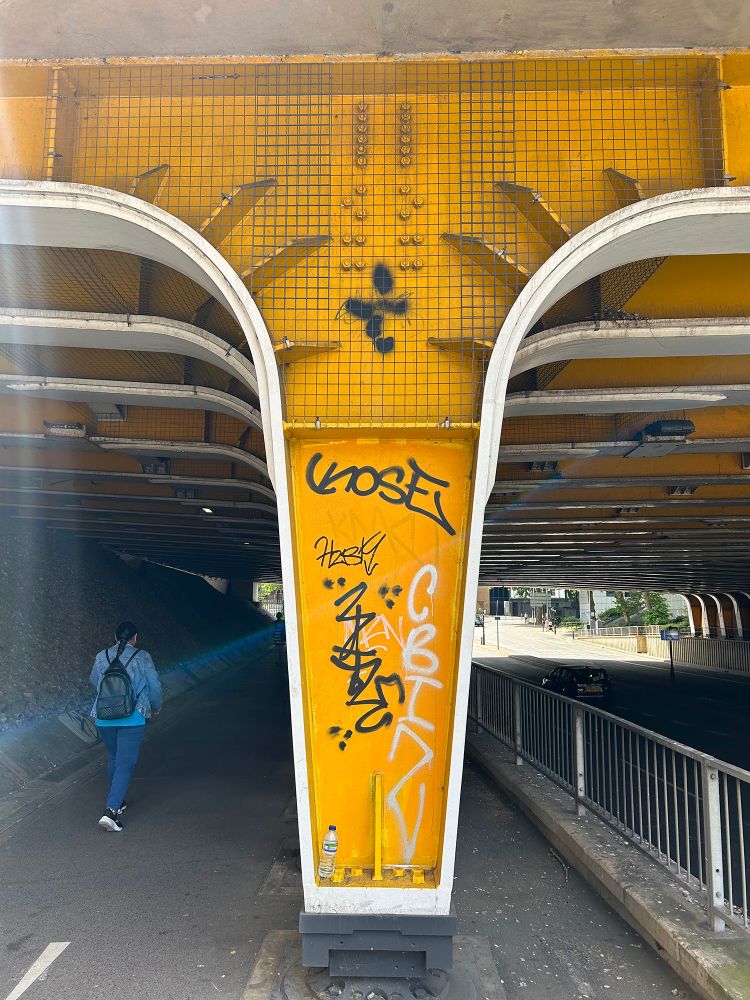 A person enters an underpass and a coincidental flare of blue light seems to illuminate their trajectory. Separating the pedestrian from  vehicular traffic are large iron supports which underpin the multiple railway tracks above. These sizeable structures are painted bright yellow and have been extensively tagged in black paint and white paint — a tasteful colour scheme if nothing else. Vastern Road, Reading, Berkshire, UK. Photo by Gearóid Burke.