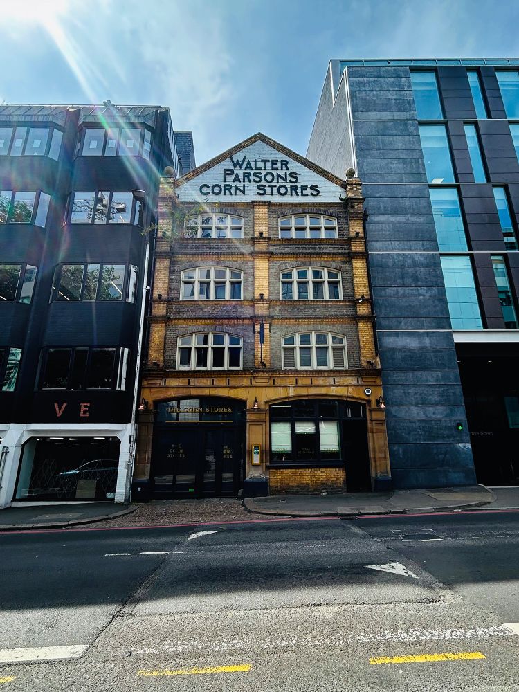 A wide-angle shot of three buildings and the sloped road in front of them. At the centre is a yellow brick, four-storey, two-bay building. This Grade II listed former warehouse was built in 1890 for Walter Parsons, corn merchants, as the legally-protected large lettering on its gable suggests. Forbury Road, Reading, Berkshire, UK. Photo by Gearóid Burke.