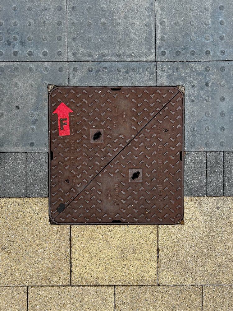 A split, square, cast-iron manhole cover is surrounding by three types of paving. It has a red, die-cut, arrow-shaped sticker with the letter F (presumably indicating flow direction) applied to the top left corner. The access cover is a dark ferrous brown colour and features a raised tactile pattern of L shapes and the name of the manufacturer, Stanley John Ltd., plus EJ Group and Python. Sentinel End, Reading, Berkshire, UK. Photo by Gearóid Burke.