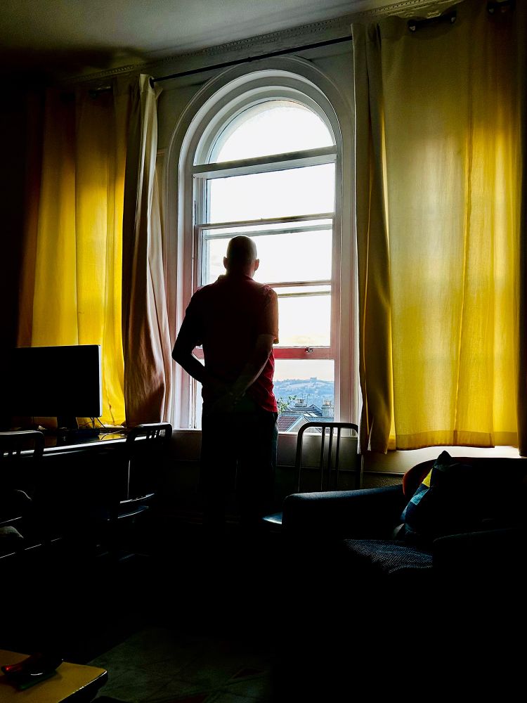 A dark interior. A man stands in silhouette, his hands behind his back. He is looking out of a window. The sash window has four horizontal panes and an arch at the top. There are two other four-pane windows, one on each side, but they are covered by curtains which allow some light through their fabric. The twilight sky beyond the glass is mostly blown out or overexposed, but in the distance is Kelston Roundhill. Bath, UK. Photo by Gearóid Burke.