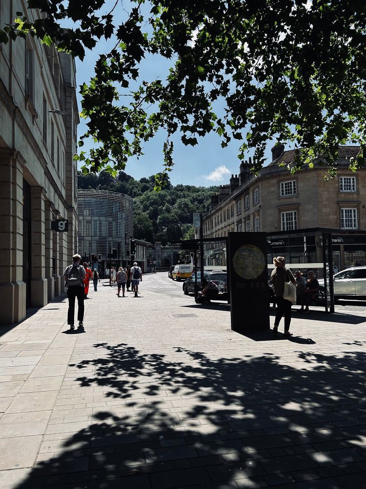 The midday sun casts strong shadows on a wide pavement. People pass a neoclassical building on the left, heading in the direction of a modern curved building. In the distance is a tree-covered hill. On the right is a dark metal monolith featuring a circular map, and people sit in the shade of a bus shelter. Southgate Street, Bath, UK. Photo by Gearóid Burke. 