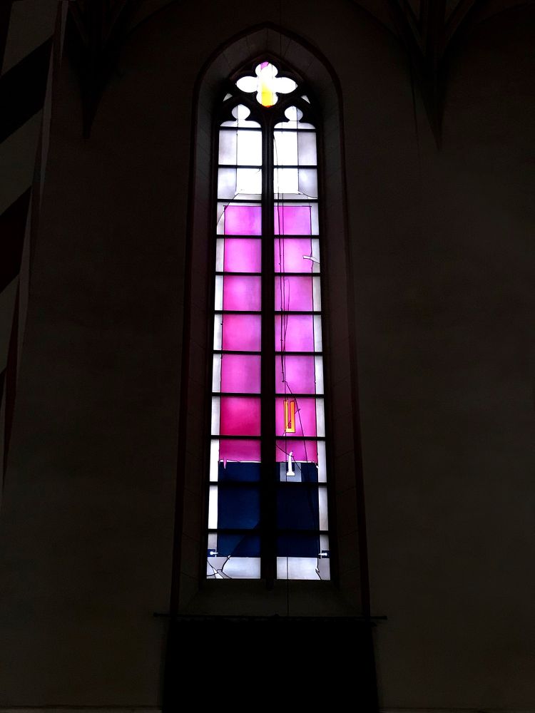 A thin Gothic church window with a quatrefoil at the top and stained glass throughout. The design is contemporary and abstract, with a large area of bright pink, and delicate details in fine lines. One of a five-window interpretation of the 22nd Psalm in colour, line, and light by Johannes Schreiter (b. 1930). St. Jacobi-Kirche, Göttingen, Lower Saxony, Germany. Photo by Gearóid Burke.