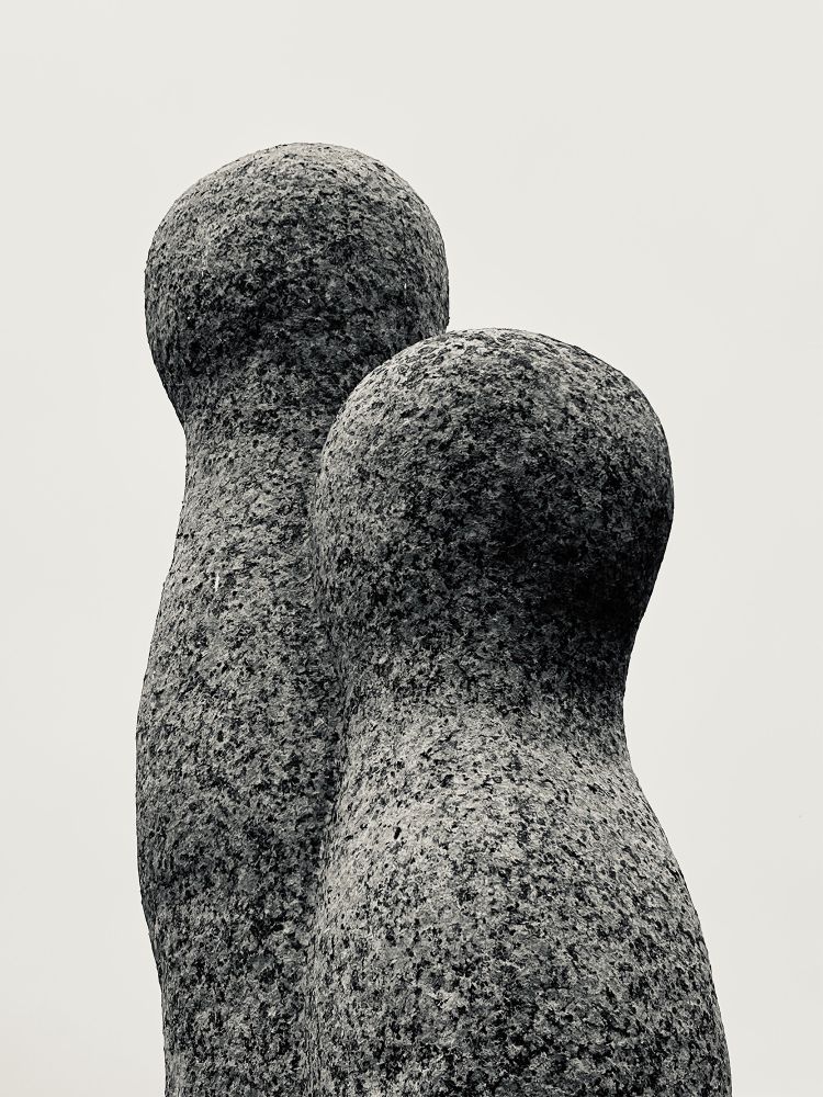 A monotone photograph of a granite sculpture featuring two figures in a rounded, minimal style. One is taller than the other. The top half of Seenu's sculpture "Éclosion" ‎⁨(2007) at Parc Des Sculptures Symposium⁩, ⁨Perros-Guirec⁩⁩, ⁨France⁩. Photo by Gearóid Burke.