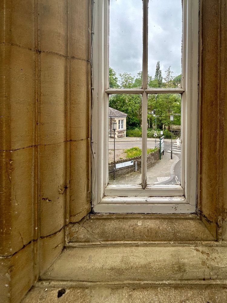 Looking through an narrow four-panel window in a yellow brick wall. A woman with white hair and a white coat crosses the white stripes of a pedestrian crossing. Platform 1, Bath Spa Railway Station, Bath, UK. Photo by Gearóid Burke.