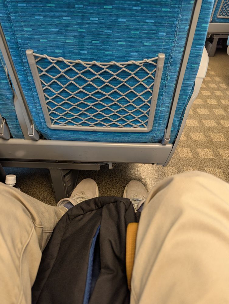 A pair of legs in khaki trousers not being  constricted by the ample legroom to the aquamarine seat in front.