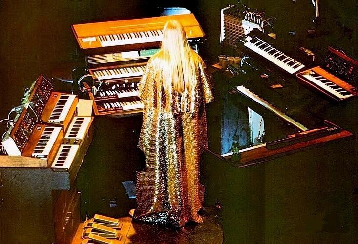 A man with long blond hair and a floor length sparkling gold cape stands with his back to us. He is surrounded by keyboards.