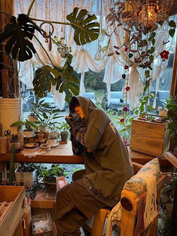  ateez Instagram post, look at him he's in baggy brown and green clothes with his hood up and sweater paws and hes in a beautiful plant store (??), with big monstera leaves and vines and little sprouts on table he's leaning against. the chair he's sitting in has a big, floral patterned pillow and weaving projects slung over the side. THIS TIME he's holding his hands in a little heart shape right next to his face, so you can now see his nose and his little pout