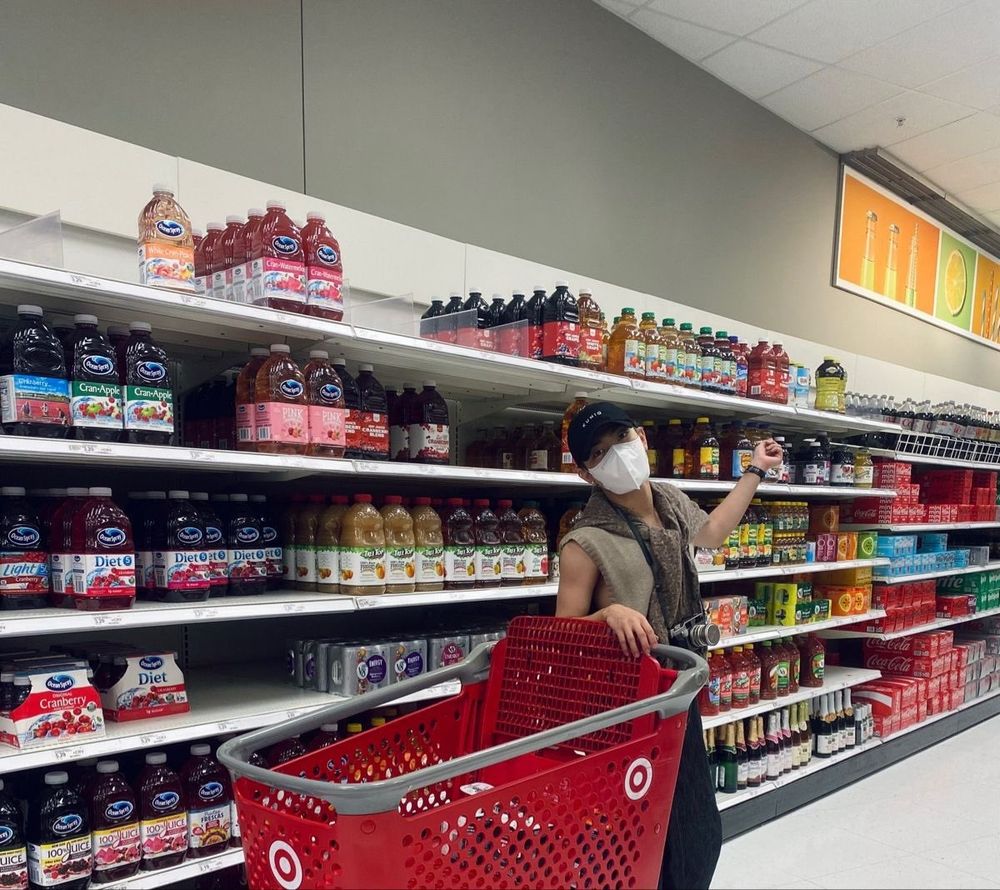 ateez hongjoong in the juice section of a target. he's wearing a white mask, a black cap, a beige sweater vest with a brown sweater (??) tied around his shoulders, and wide black pants. he also has a camera around his neck. he's leaning against one of those massive target shopping carts and pointing behind him at the juice like "get a load of this guy," the guy in question being the juice??? maybe!