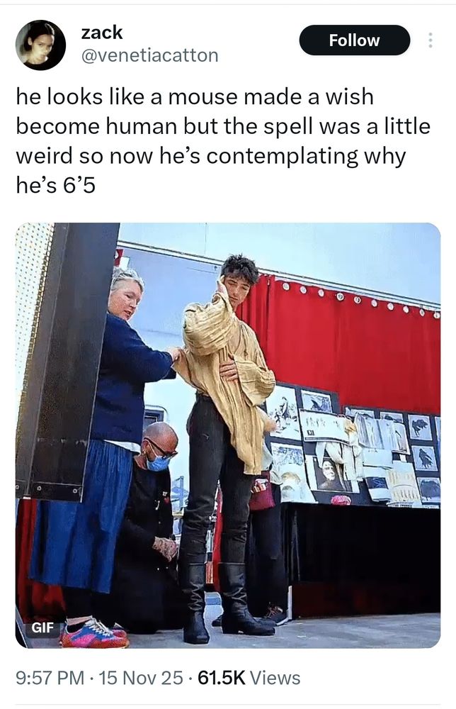 screenshot of a tweet by user @venetiacatton 

the image is of Jacob Elordi being dressed for Frankenstein, he's in a big, baggy, dark cream, peasant adjacent blouse, skinny cut black pants, and black boots with a slight heel. one of his hands is resting over his torso, the other is scratching the side of his head. he has a sleepy expression on his face. he is towering over the three staff members helping him get into costume. 

the text of the tweet says, "he looks like a mouse made a wish to become human but the spell was a little weird so now he's contemplating why he's 6'5"