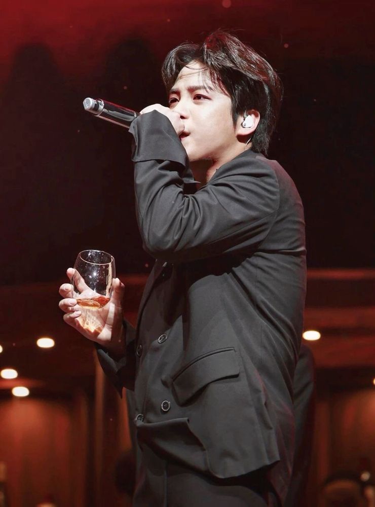 ateez jongho during arriba, he's wearing a black suit and holding a glass of what they want us to think is brandy or whiskey or something. He is holding a microphone up to his mouth with the other hand. his dark brown hair is windswept 
