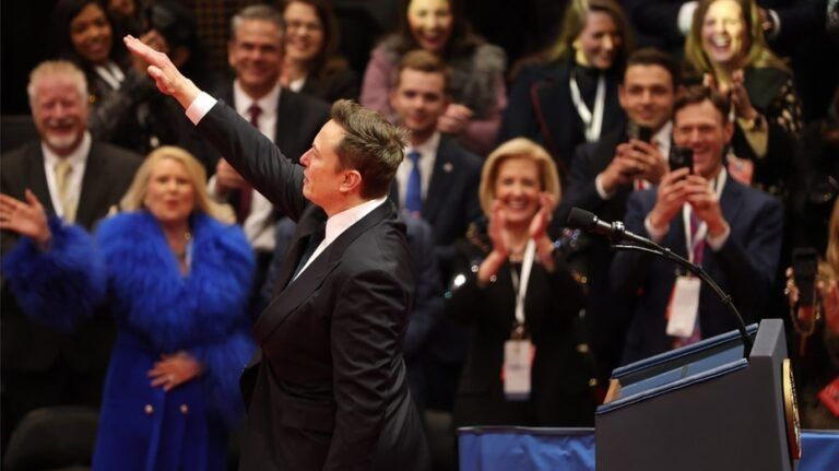 The crowd of Nazis looks on with glee as the Nazi on stage performs a Sieg Heil.