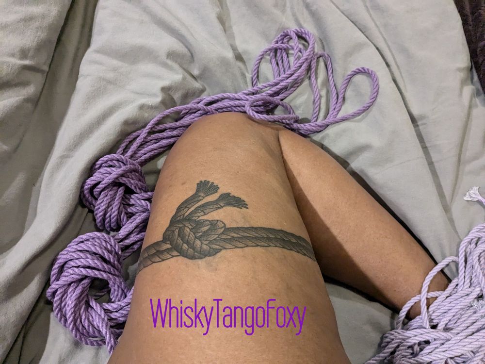A Black woman shows off a black ink tattoo on her right thigh, resting in a bed of orchid purple hemp rope. The tattoo is a sheet bend rope join and is placed where the self tyer can balance in a single rope, single column, hip-key style suspension