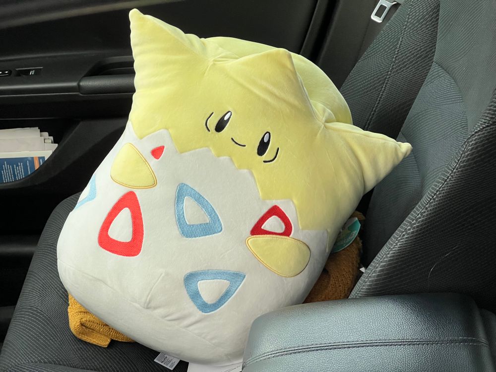 A Togepi squishmellow (overstuffed plushie) about 16 inches (40cm) tall, riding shotgun

Togepi is innocent and joyful and lucky, but always looks slightly stressed out

Togepi (トゲピー, spiky-peep) is a fairy-type Pokémon, #0175 in the Pokédex, and is associated with the trainer Misty. It is a spiky-headed yellow baby bird who has only partially hatched. Its hands and feet poke out from the shell, and is otherwise covered below the head. The shell is white with red and blue rounded triangular rings for spots