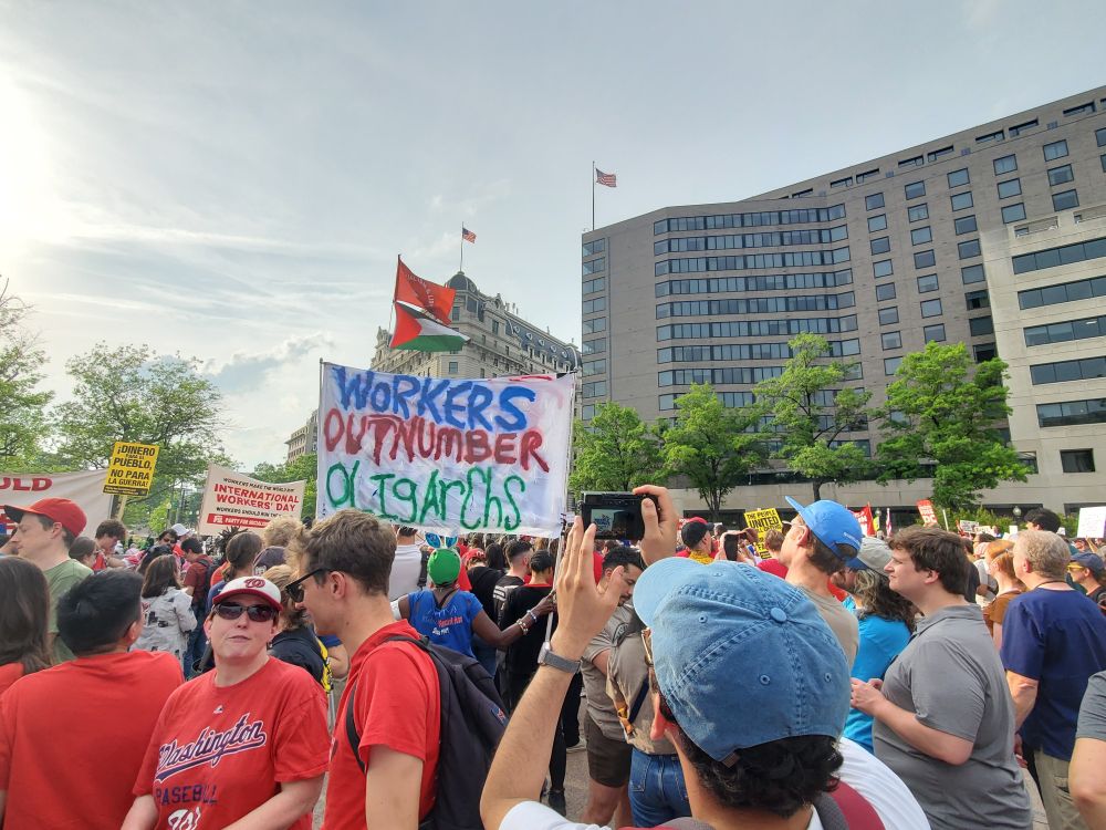 A crowd of workers gathers at the protest and someone holds up a banner that reads workers outnumber oligarchs.