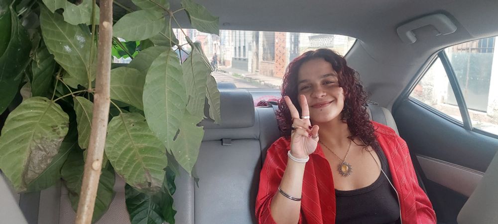 uma moça de cabelos cacheados, camiseta preta e blusa vermelha no banco de trás de um carro com uma árvore de ipê ao seu lado.