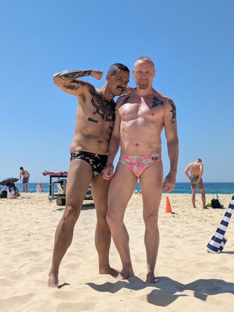 Juan and Timo on the beach. Juan is in a camo print speedo, Timo’s pink speedo has cockatoos 