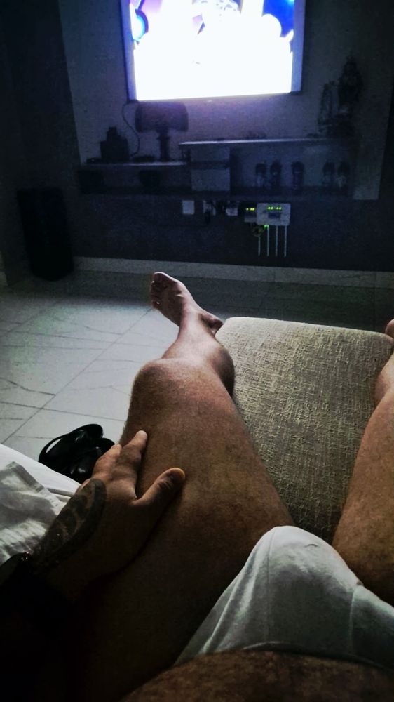 Pessoa deitada em um sofá, com as pernas esticadas em direção à frente. A mão esquerda repousa sobre a coxa, mostrando parte de uma tatuagem no braço. Veste roupa clara. Ao fundo, uma televisão ligada ilumina a sala escura; abaixo dela, há prateleiras com objetos decorativos e equipamentos eletrônicos com luzes verdes acesas.