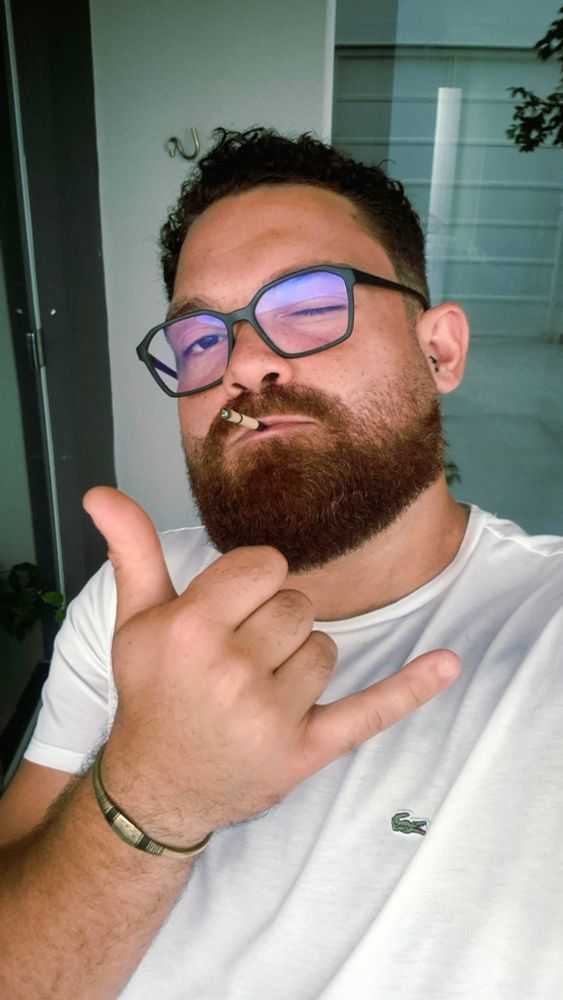 Homem de barba castanha-avermelhada, cabelos cacheados curtos e óculos de armação escura com lentes azuladas tira uma selfie em ambiente interno. Veste camiseta branca da Lacoste, usa pulseira metálica no pulso esquerdo e segura um cigarro fino entre os lábios. Está piscando com o olho direito e fazendo o gesto de hangloose com a mão esquerda, com o polegar e o dedo mínimo estendidos. Ao fundo, há uma parede branca, uma porta de vidro e reflexos de vegetação.