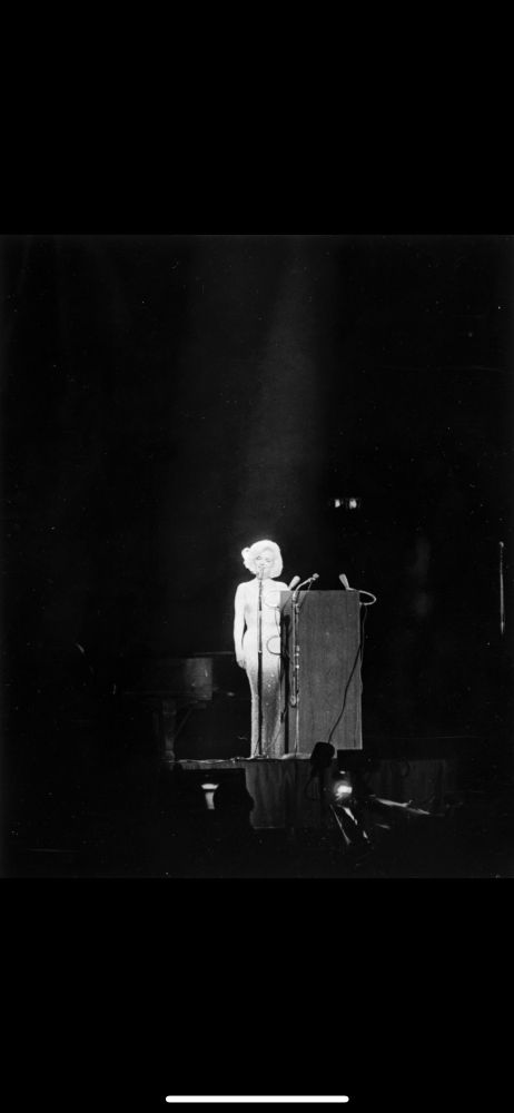 Black and white photo of Marilyn Monroe on stage at Madison Square Garden, the night she sang Happy Birthday Mr President in 1962 to JFK in her Sexy Baby voice.  The framing of the wide shot puts her full
body at the size of a 1/9 segment right in the centre.  All other eight segments are black, and a single spotlight shines down onto her, and then bounces right off her, her platinum hair, the northern European skin of a dedicated Californian tan-avoider, and the infamous diamanté-studded gown, making her the only illumination in the shot/stage/theatre.