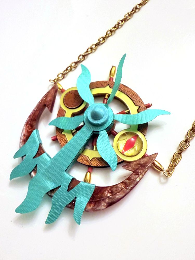 Photo of a necklace pendant made from laser cut acrylic, veneered wood and ribbons with beadwork embellishments. The piece resembles Dhelmise, a Pokémon which combines visual elements from a nautical steering wheel, a ship's anchor and kelp.