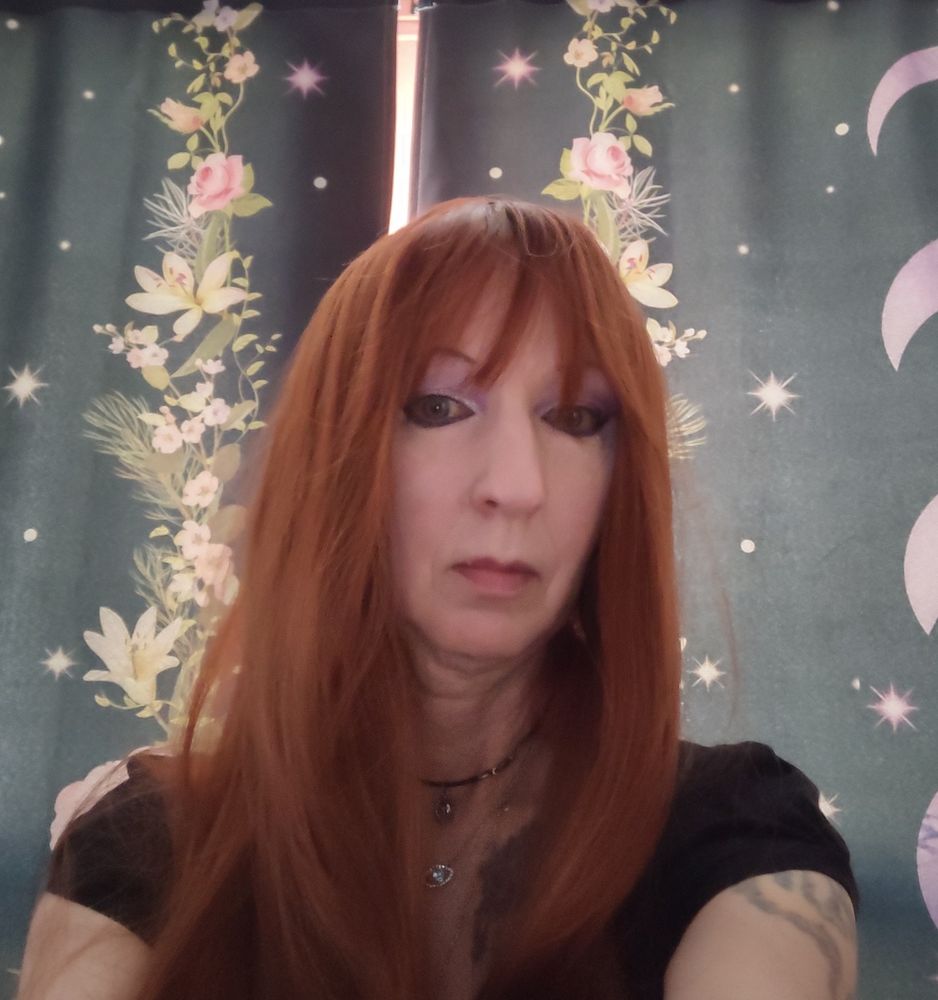 Person wears a long auburn wig with longish curtain bangs in a classic late sixties, early 70's L.A. style. Eyes are smokey and lips tinted a light wine color.  In the background are black curtains with a floral and moon pattern.