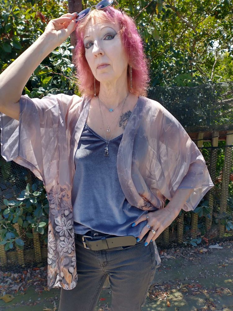 Person with shaggy redpink hair with orange bangs raises sunglassed onto top of head. They are wearing a diaphanous grey short kimono with peach tones, and white, grey & peach flowers decorating the bottom. This is matched with a grey velvet tank top, faded black jeans and a black chunky belt, and silver jewelry. 