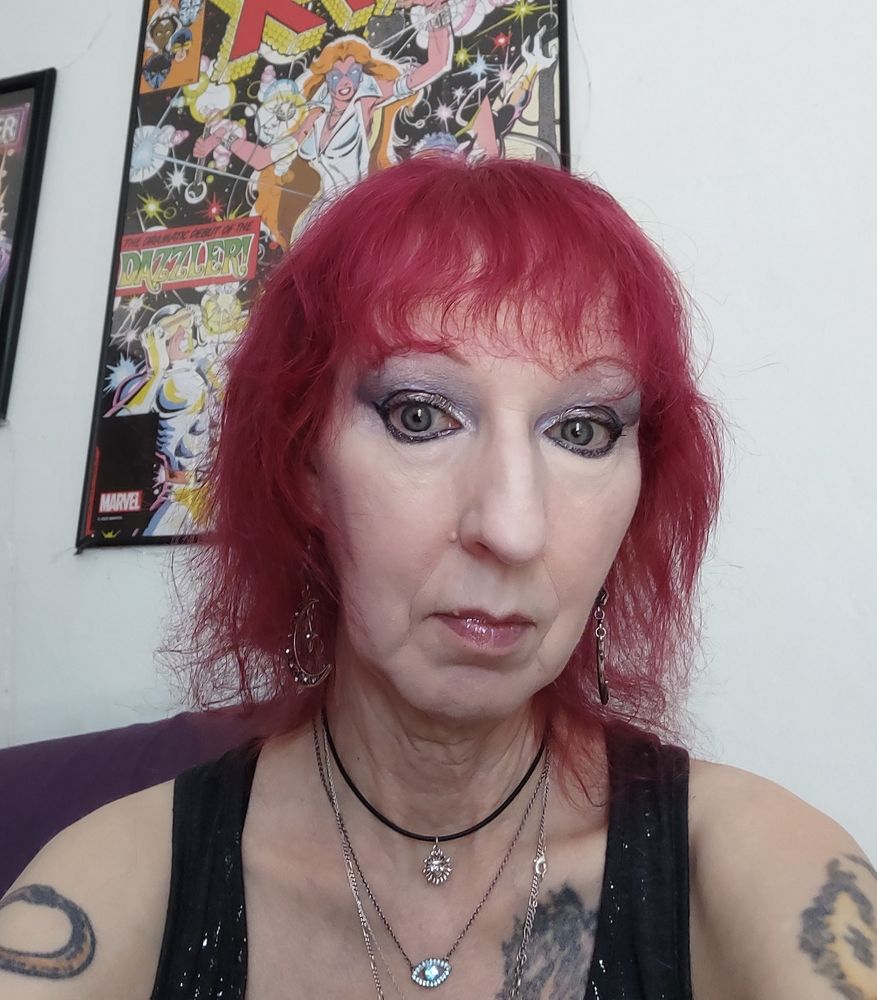 Photo of person with a fuchsia pink shag haircut with wavy bangs, with smokey painted eyes. they have multiple tattoos partially visible - a phoenix on arm and shoulder, an infinity ouroboros on the other arm, & wearing a grey tank top shot through with silver, multiple necklaces including a small sun symbol choker, and an "evil eye" pendant. They wear a somewhat sad expression of thoughts far away. Behind them is a poster of Dazzler from The X-Men.