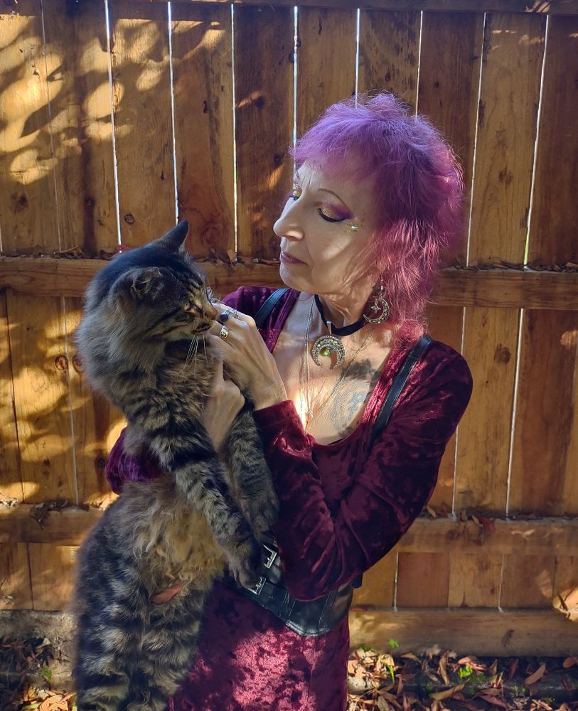 Person with pink and purpleish hair cut in a shag wears a burgundy velvet dress and silver moon jewelry, while holding a large fluffy brown tabby cat. They stand in a leaf strew yard in front of a sun-dappled wooden fence.
