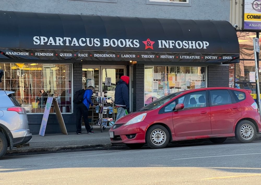 Street view with black coloured awning  showing the words “Spartacus Books Infoshop” with a red star and cars parked in front of store front windows. 
