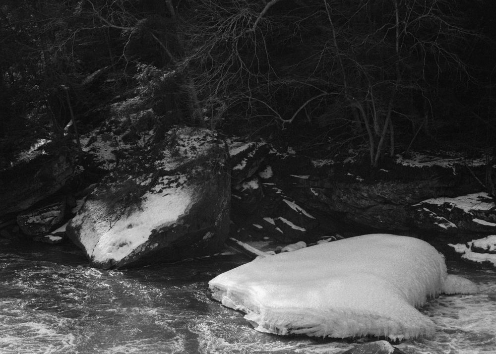A black and white photo of a river rushing past some large snow-covered rock slabs. One is slanting from the opposite riverbank into the water; the other is flat and protruding into the river, covered in about a foot of snow. The forest on the other side is underexposed and the details are crushed into black.