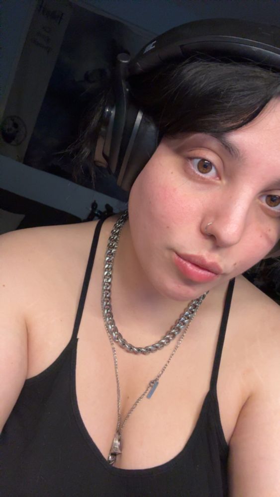 Me with big black headphones, silver jewelry, and a black tank top with Rosie cheeks and no make up on my face. It’s a selfie. I have brown eyes.