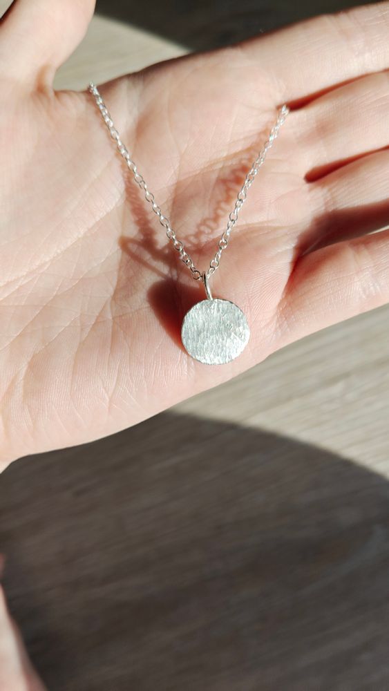 A silver chain in the sunlight with a silver pendant with a lot of texture, looking kind of some bark texture. Much shiny such wow.