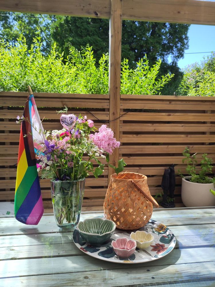 picture of a vase with wild flowers and the progress flag on a wooden table, with a wooden fence and a hedge behind it.