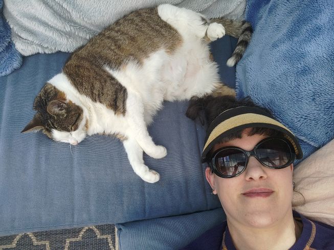 picture from above, of a woman in sunglasses lying next to a sleeping cat. 