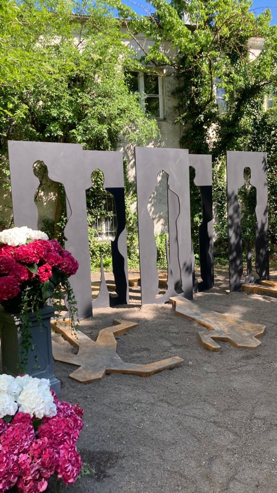Skulptur auf 5 Metallplatten, aus denen jeweils die Silhouette eines Menschen herausgetrennt wurden. Am Boden liegen menschliche Silhouetten davor oder dahinter. Daneben Blumengestecke. 