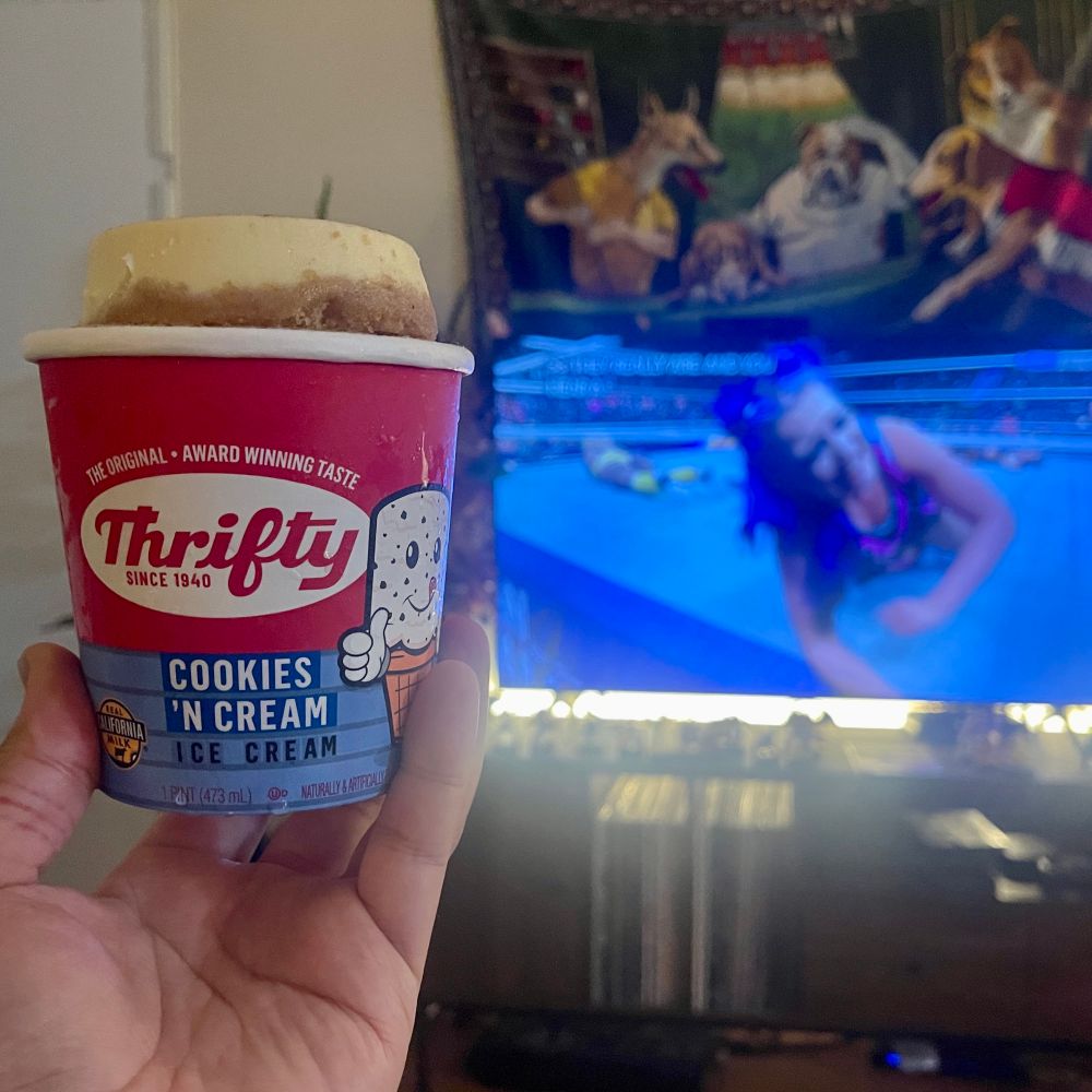 a hand holding a pint of Thrifty Cookies ‘s Cream ice cream topped with a tiny cheesecake while AEW plays in the background
