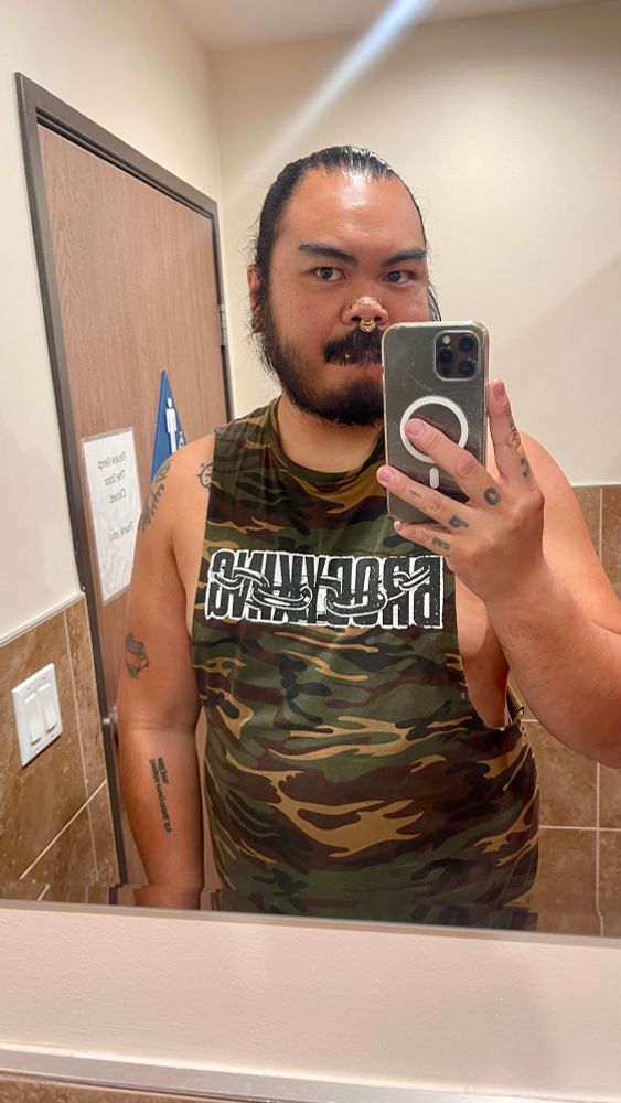user jaddicusfinch taking a bathroom mirror selfie wearing an edited camouflage Brody King shirt that is cut so deep it is barely covering their nipples. unfortunately, they’re unconsciously doing bearface