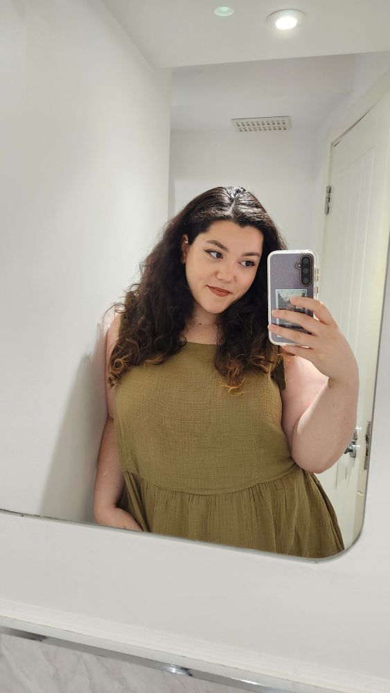 Becca smiles, looking at the phone in her hand in a mirror selfie. A Polaroid of Nate in her phone case peeps through her fingers. 