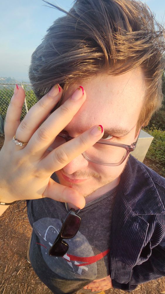 Another picture on 0.6x zoom. Beccas hand covers Nates exasperated face to show off new engagement ring. It is a gold ring with 3 diamonds, and was formerly Beccas grandmother's ring.