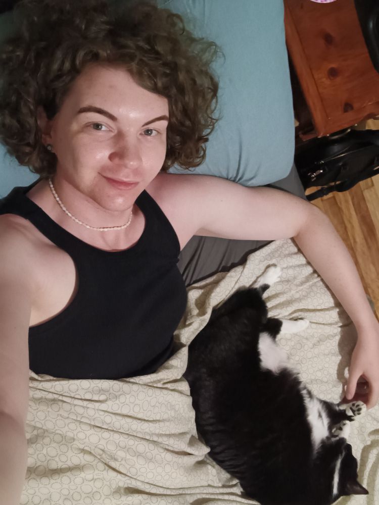 Me in bed in a black dress and white necklace spooning my chunky tuxedo cat