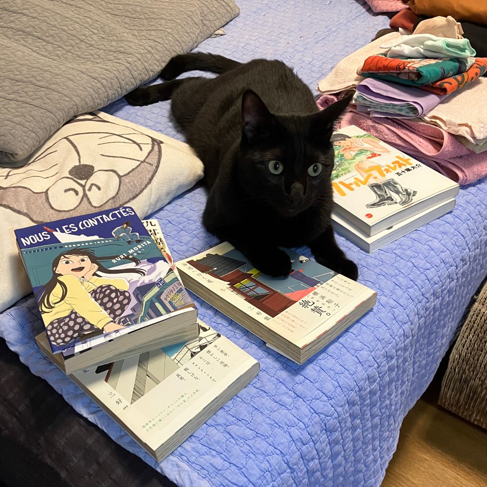 『海辺へ行く道　冬』の上に前足をのせる黒猫の写真。
黒猫のまわりにはほかにもたくさん漫画の本が散らばっている。