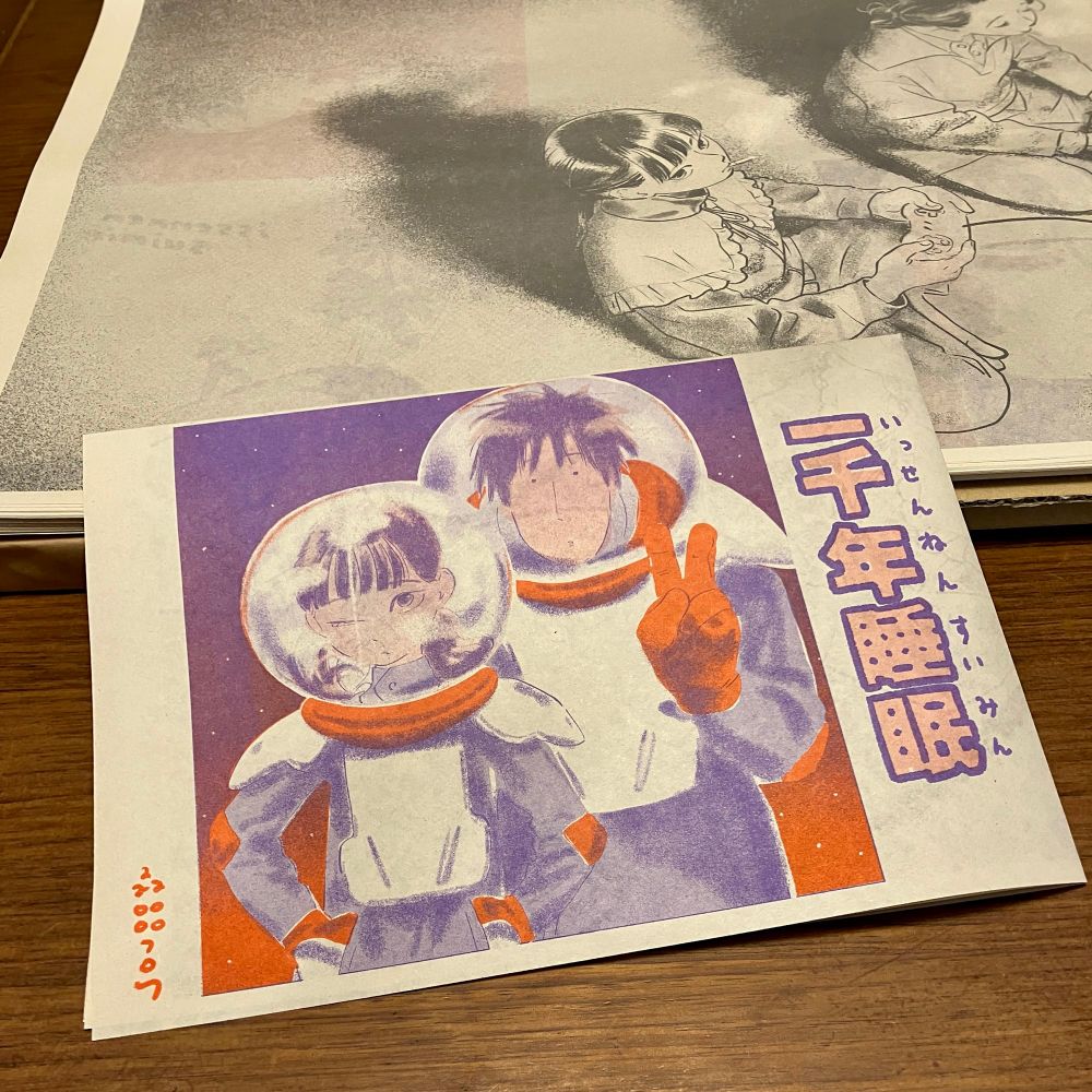 リソグラフ印刷物の画像。
木のテーブルの上に、四つ折りにされたリソグラフ印刷紙と、その下に広げたリソグラフ印刷紙が置いてある。
四つ折りの方は「一千年睡眠」「森田るり」の文字。『我らコンタクティ』の主人公、椎ノ木カナエと中平かずきが宇宙服を着てこちらを見ているイラスト。
広げた方にも同じく椎ノ木カナエと、その奥に梨穂子という同マンガの登場人物が、あぐらをかいて座っているイラスト。