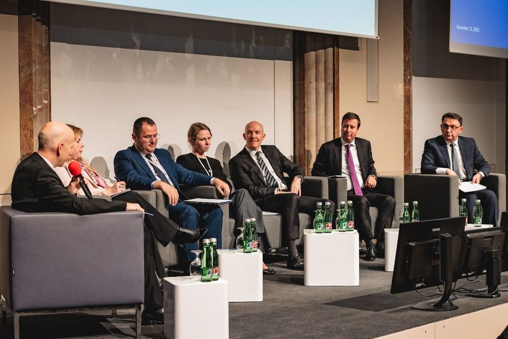 The image shows a panel of six people in business attire sitting on a stage, engaged in a discussion or presentation at what appears to be a professional conference or event.