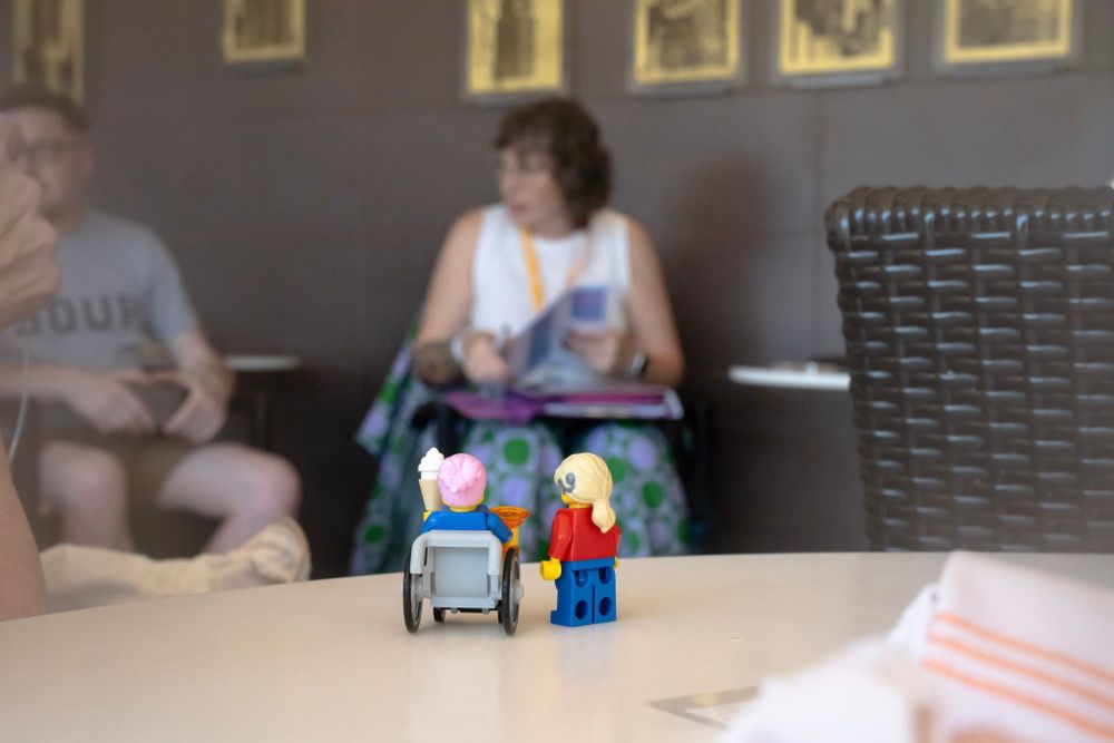 Two LEGOs are in the foreground. One has short pink hair and is in a grey wheelchair holding an ice cream cone. The other wears a red shirt and blue pants, has long blonde hair and a hearing aid. They are facing away from the camera, as if taking part in the meet up